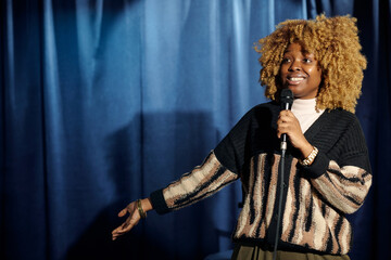 Young smiling female stand up comedian speaking in microphone during stage performance while...