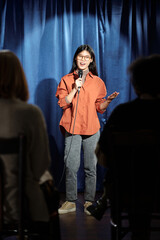 Young comedian of stand up club speaking in microphone while standing on stage with blue curtains...