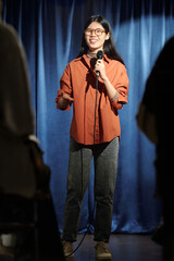 Young smiling comedian in casualwear standing on stage with blue curtains and speaking in...
