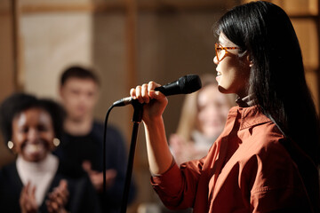 Young female comedian in eyeglasses standing in front of camera against audience and speaking in...