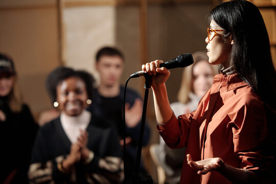Young comedian of stand up show standing against audience and pronouncing monologue in microphone during performance