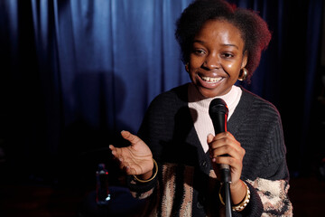 Happy young female comedian of modern stand up club holding microphone while speaking in front of...