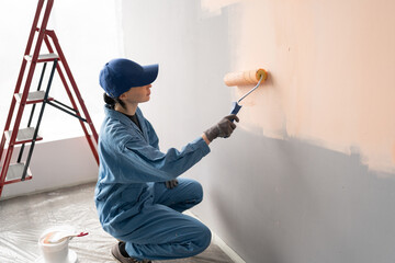above view of female painter in overalls with cap and gloves painting a wall with paint roller,...