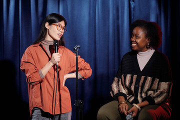 Young Asian female comedian speaking in microphone and looking at African American artist sitting against blue curtains on stage - obrazy, fototapety, plakaty