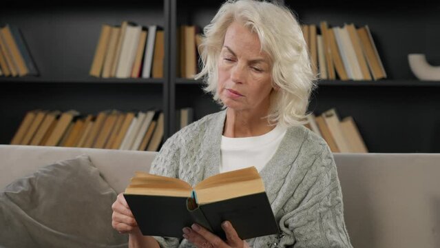 Senior smiling woman reading book. Retired female bookworm bibliophile book lover enjoys interesting story sitting on couch at home. University professor resting concentrated, leisure weekend concept.