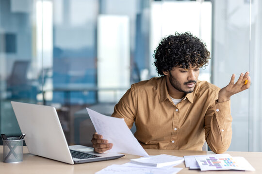 Clueless diverse male checking printed documents on desktop during working day in modern office interior. Confused man in beige shirt looking at statistic charts at cabinet with glassy walls.