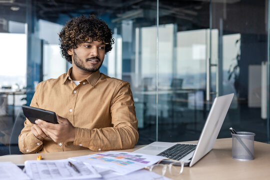 Calm latino guy looking at laptop screen while sitting by desktop with scattered charts and documents. Attentive accountant holding calculator and checking data in electronic company expenses report.