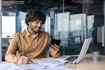 Smiling hispanic man looking at calculator while filling in printed table next to pc in modern office. Careless remote financier using electronic gadget for reducing time for data calculation.