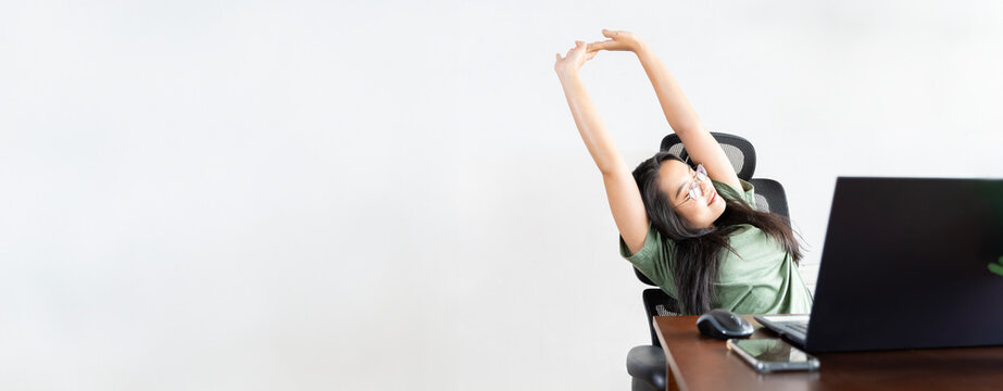 Happy relaxed young woman sitting in her kitchen with laptop in front of her, stretching her arms above her head and looking out the window.