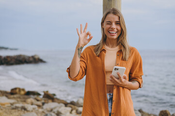Young smiling happy fun woman she wears orange shirt casual clothes hold in hand use mobile cell...