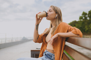 Side view young calm woman wears orange shirt casual clothes listen music in earphones drink coffee...