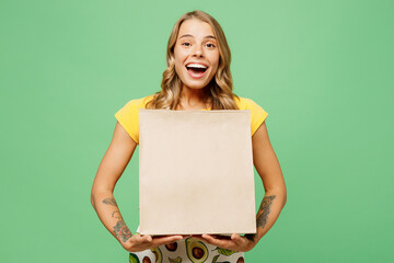 Young surprised housewife housekeeper chef cook baker woman wear apron yellow t-shirt hold in hand paper craft brown blank bag isolated on plain pastel green background studio. Cooking food concept.