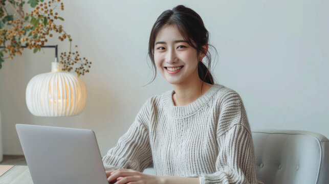 A cheerful young woman smiles while using a laptop, seated at a minimalistic desk with soft lighting.