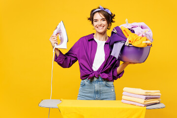 Young smiling woman she wear purple casual shirt do housework tidy up ironing clean clothes on board hold basin with laundry isolated on plain yellow background studio portrait. Housekeeping concept.