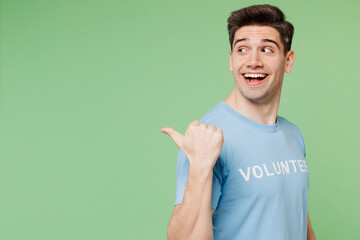 Young smiling man wears blue t-shirt white title volunteer point thumb finger aside on area isolated on plain pastel green background. Voluntary free team work assistance help charity grace concept.