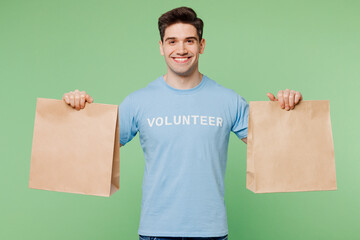 Young man wears blue t-shirt white title volunteer hold in hand two paper brown blank bags delivery isolated on plain pastel green background Voluntary free work assistance help charity grace concept