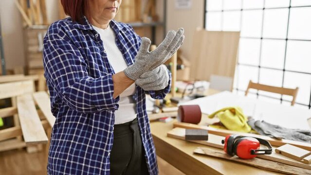 A mature woman wearing gloves in a carpentry workshop filled with tools and wood.
