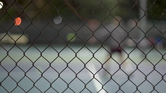 Cinematic shot of tennis player playing tennis on court at night. Focus on mesh fence, player blurred in the background.