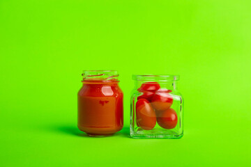 A small glass bottle of fresh tomatoes juice and a small square jar of cherry tomatoes on a green background. Delicious fresh vegetables. 