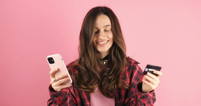 Young woman show mobile and card using mobile cell phone hold in hand credit bank card do online shopping book tour isolated on pink background. Wow surprised girl look at phone, show credit card.