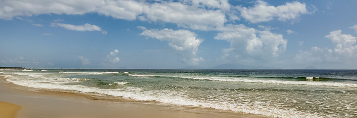 Tranquil tropical beach panorama with gentle waves, clear blue sky, and distant mountains, ideal for travel and tourism backgrounds with copy space
