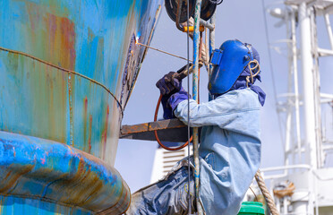 Welder rappelling with ladder to welding metal hull surface of the old fishing boat during maintenance and improvement work in shipyard area - obrazy, fototapety, plakaty