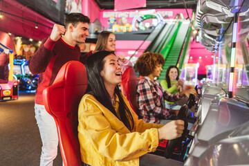 Asian woman playing with her multiethnic friends in racing games in an arcade game center while her friends celebrate and cheer