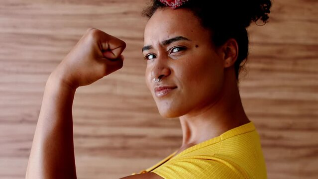 Bold African American Lady in Yellow Displaying Power – Muscular Pose and Direct Gaze, Symbol of Determination 'We Can Do It'. Woman flexing muscle with serious expression