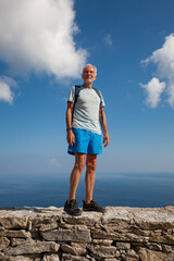 Caucasian senior male standing on the stone wall with blue sea and cloudy sky in a background (Amorgos, Cyclades/Greece)