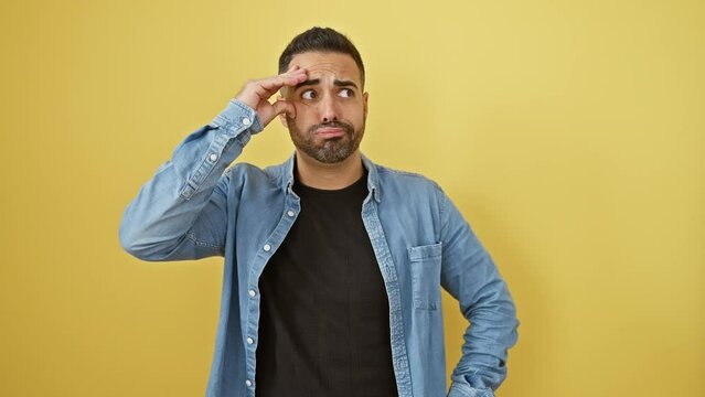 Worried young hispanic man in denim shirt stressing over a crisis, hand on head, standing against isolated yellow background.