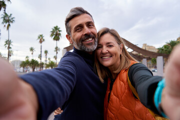 Cheerful selfie of a middle age caucasian couple looking to the camera smiling, using phone to take...