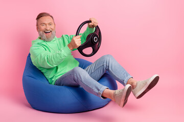 Full length photo of handsome senior man sit bean bag driving hold rudder dressed stylish green garment isolated on pink color background