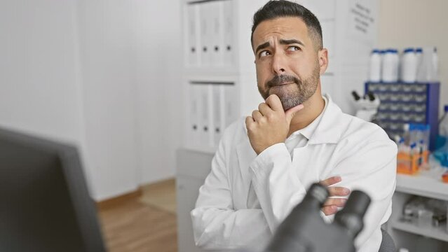 Pensive young hispanic scientist, finger on chin, deep in thought at lab computer, brewing wonder with a dash of doubt.
