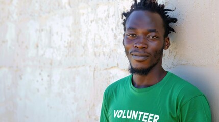 Young man with dreadlocks wearing a green volunteer t-shirt leaning against a textured wall.