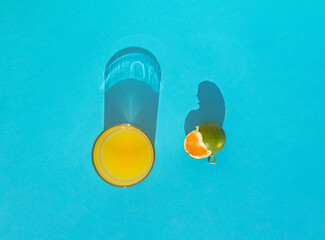 Creative concept with orange juice and green tangerine on sunny day. Fruit, drink and shadows on blue background. Flat lay.
