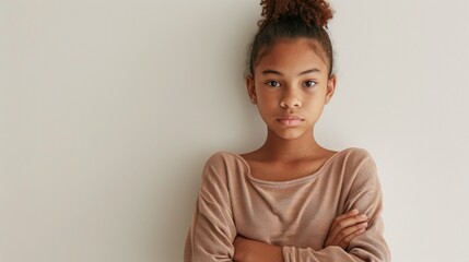 Young girl with curly hair wearing a beige top standing against a plain wall with her arms crossed looking directly at the camera.
