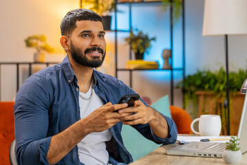 Positive Indian man using smartphone while staying in modern workplace at home. Efficient Hispanic...