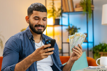Rich young Indian bearded man freelancer using smartphone and counting money cash, calculate...