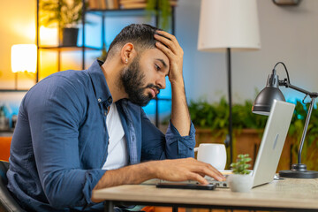 Bored sleepy young Indian bearded man working on laptop computer, leaning on hand at home office. Exhausted tired Arabian male freelancer workaholic man. Employment, occupation, workless.