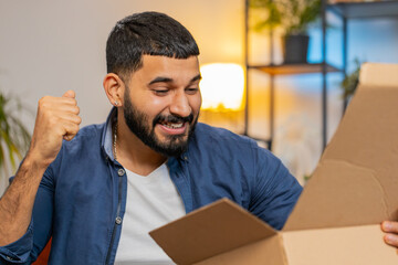 Excited young Indian man unpacking delivery parcel sitting on sofa in living room. Smiling Arabian guy shopper online shop customer clenching fists receiving purchase by fast postal shipping courier.