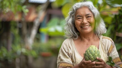 A smiling older woman with gray hair wearing a patterned top holding a large green artichoke setting with blurred greenery and a wooden structure in the background.