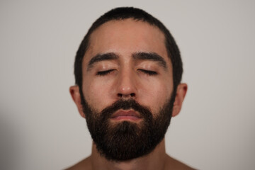 Obraz premium A closeup studio portrait of a man with closed eyes, posing shirtless against a white background. Soft lighting draws attention to his dark beard, strong eyebrows, and the hairline of his trim hair.