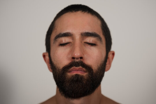 A closeup studio portrait of a man with closed eyes, posing shirtless against a white background. Soft lighting draws attention to his dark beard, strong eyebrows, and the hairline of his trim hair.