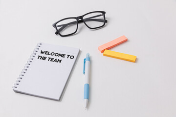 A white desk setup showcasing a notebook with Welcome to the team written on it, blue pen, black glasses, highlighters, symbolizing job opportunity and office work environment