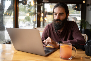 Pensive handsome male blogger with long beard working uses social networks watch multimedia files sitting in cafe with equipment and laptop computer - obrazy, fototapety, plakaty