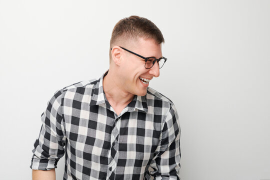 Cheerful young man in glasses and plaid shirt laughing against a white background.