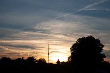 The sun gracefully sets, casting a warm glow behind a tree and a building in the evening sky.
