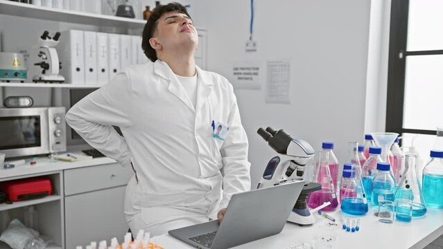 A man in a white lab coat experiences back pain in a modern laboratory with a microscope and laptop. - Powered by Adobe