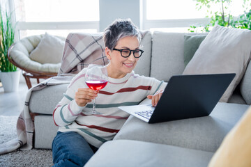 Portrait of positive pretty aged lady sit carpet floor enjoy wine glass use wireless laptop modern...