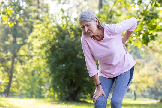 Active senior woman in sportswear touching her knee in discomfort while exercising in a sunlit park, depicting health issues.